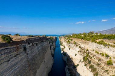 Korint Kanalı 'nın yüksek açılı manzarası. Dik, güneşli kireçtaşı duvarları ve uzak mavi denize doğru uzanan dar bir su yolu. Nadir yeşillik ve berrak bir gökyüzü çarpıcı, açık bir manzara yaratır..