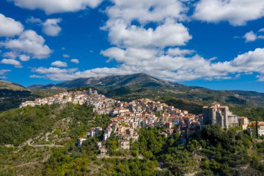 İtalya 'nın Rivello kentinin geniş hava manzarası, dağınık bulutlarla dolu bir gökyüzünün altında dramatik dağlar tarafından desteklenen yeşil bir yamacın tepesinde kümelenmiş terracotta çatıları. Kıtır gün ışığı, manzaranın zengin renklerini ve dokularını güçlendirir..