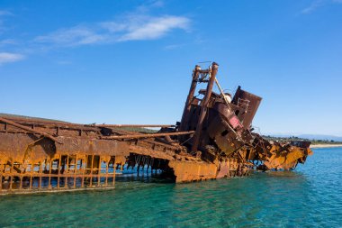 Yunanistan 'ın Gythio kentindeki kumlu bir sahil şeridi yakınlarında turkuaz sudan yükselen pürüzlü, paslı metalle Dimitrios' un gemi enkazının yakın görüntüsü. Parlak güneş ışığı, berrak mavi gökyüzüne karşı çürümüş dokuları ve canlı renkleri vurgular.
