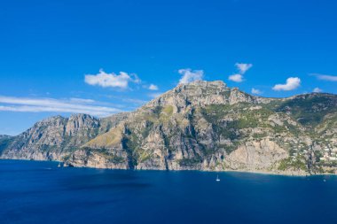 Saf kayalık dağlar berrak bir gökyüzü altında parlak mavi sulardan yükseliyor, dağınık beyaz bulutlar ve engebeli kıyı şeridini aydınlatan güneş ışığı. Sahne dramatik manzarayı ve İtalyan kıyı bölgesinin tipik doğal dokularını yakalar..