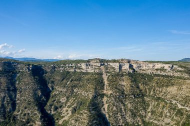 Navacelles Sirki 'nin derin, yeşil kaplı kanyonunda yükselen engebeli kireçtaşından bir yamaç manzarası. Parlak güneş ışığı, açık mavi gökyüzünün altındaki kayalık dokuları ve seyrek bitkileri güçlendirir..