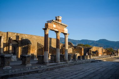 Flütlü sütunları olan antik Roma kemeri, sabah güneşiyle aydınlanan İtalya 'nın Pompeii kentindeki sivil forumda duruyor. Yıpranmış tuğla ve taş detaylar açık mavi bir gökyüzü ve uzak bir dağ zeminine karşı dizilmiştir..
