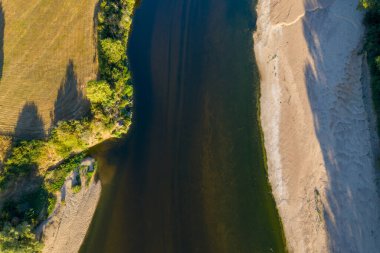 Geniş kumlu bir sahil ve yeşil bitki örtüsüyle çevrili dolambaçlı bir nehrin havadan görünüşü. Altın tarlalar uzun gölgeler bırakıyor. Resim, doğal dokuları, zıt renkleri ve yazın kırsal bir nehir manzarasının sakin atmosferini ön plana çıkarıyor..