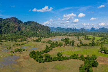 Geniş pirinç tarlaları ve dağınık ağaçlar Laos kırsalındaki canlı mavi gökyüzünün altında dramatik kireçtaşı tepelerine doğru uzanır. Manzara doğal desenler, yemyeşil yeşillik ve sakin kırsal manzaraya sahiptir..