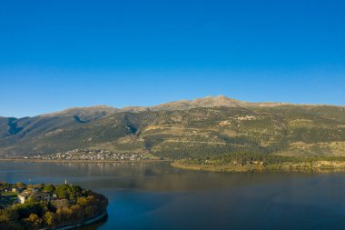 Yumuşak sabah ışığı, Ioannina Gölü 'nün üzerindeki orman yamaçlarını ve kayalık tepeleri aydınlatır. Pindus dağlarının eteklerini yansıtan sakin su. Sahne, Yunanistan 'ın Epirus kentindeki geniş mavi gökyüzü ve doğal dokuları yakalıyor.