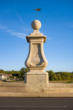 Süslü bir taşın, üzerinde küresel bir final ve metal rüzgar gülü bulunan, canlı, mavi bir gökyüzüne yakın plan. Sıcak güneş ışığı, arka planda görünen uzak ağaçlar ve çatılarla desenli yüzeyi vurgular.