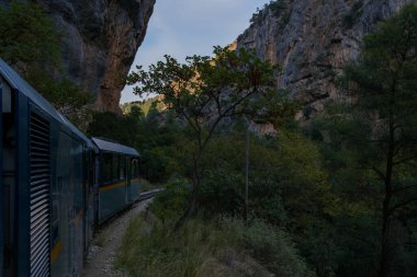 Yunanistan 'ın Kalavrita kenti yakınlarındaki yoğun yeşillik ve dik kayalıklarla çevrili kayalık bir vadide dar bir tren esiyor. Yumuşak akşam ışığı, Mora dağlarındaki engebeli manzarayı ve eşsiz demiryolu yolculuğunu vurgular..