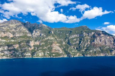 Yeşil bitki örtüsüyle kaplı kayalık dağlar beyaz bulutlarla dağılmış bir gökyüzünün altındaki canlı mavi sulardan yükseliyor. Gıcır gıcır gün ışığı, bu İtalyan kıyı manzarasının doğal dokularını ve dramatik hatlarını vurgular..