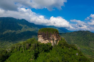 Yoğun yeşil ormandan, yuvarlanan tepelerle çevrili ve parlak mavi bir gökyüzünün altında dağınık bulutlar altında canlı bitki örtüsüyle kaplı dramatik bir düz kaya oluşumu ortaya çıkar. Manzara, vahşi dokuları ve yeşillikleri yakalar.