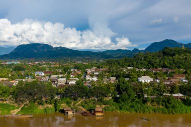 Luang Prabang, Laos 'un havadan görünüşü, çamurlu Mekong Nehri boyunca yemyeşil verimli tropikal çatıların arasına kurulmuş geleneksel çatıları gösteriyor. Dramatik kümülüs bulutları orman dağlarının üzerinden yükselir, parlak gün ışığı altında canlı ve huzurlu bir manzara yaratır..