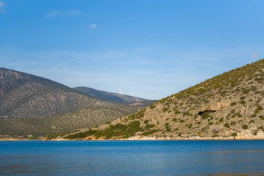 Distant wind turbines line the ridge of sunlit hills above a calm blue bay near Kilada, Greece. Rocky slopes dotted with low shrubs create a tranquil Mediterranean landscape under a clear sky.