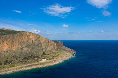 Monemvasia 'nın dik turuncu kayalıkları geniş mavi gökyüzünün altında dolambaçlı bir kıyı yoluna ve canlı turkuaz denize bakar. Dramatik manzara, kayalık dokuları ve doğal karşıtlıkları vurgulayarak güneş ışığıyla yıkanır..