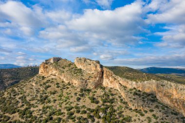 Düşük çalılarla benekli yuvarlanan tepelerin üzerinde belirgin bir kireçtaşı tepesi yükseliyor, dağınık bulutlar ve canlı mavi tonlarla dolu bir gökyüzünün altında. Engebeli arazi ve doğal dokular açık bir vahşi doğa hissi uyandırıyor..