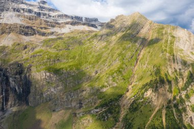 Pireneler 'deki parçalı bulutlu bir gökyüzünün altında yeşil çimen çizgileri ve açıkta kalmış kaya katmanları olan dağ uçurumları. Öğleden sonra güneş ışığı dramatik dokuları ve keskin jeolojik özellikleri vurguluyor.