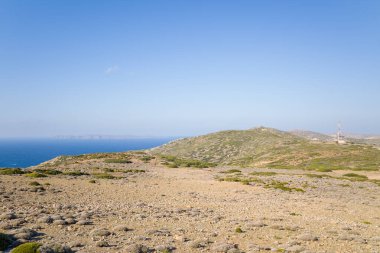 Arid rocky terrain stretches toward low green hills and the deep blue Mediterranean Sea under a clear sky in Crete. Sparse vegetation and distant islands create an open, tranquil landscape.