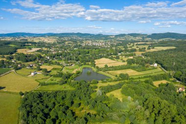 Yama alanları, sık ormanlık alan ve Saint-Honore-les-Bains yakınlarındaki dağınık kırsal evler ile çevrili yansıtıcı bir gölet gösteren canlı hava manzarası dağınık mavi bulutlar altında.