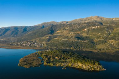 Uzaktan yükselen Pindus dağlarının engebeli yamaçlarına sahip derin mavi göl sularıyla çevrili, ağaçlarla kaplı Ioannina adasının havadan görünüşü. Sahnede yemyeşil, doğal dokular ve Epirus 'ta berrak sabah ışığı yer alıyor.