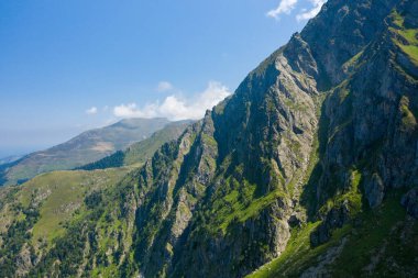 Dik kayalar, açık mavi bir gökyüzünün altında, Pireneler 'in yeşil yamaçlarında parlak güneş ışığıyla yıkanmış yeşil yamaçların üzerinde keskin bir şekilde yükselir. Engebeli taş ve yumuşak bitki örtüsünün çarpıcı kontrastı çarpıcı bir doğal manzara yaratır..