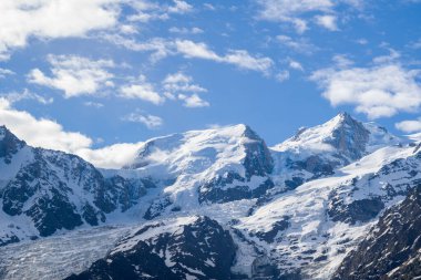 Bright midday light highlights the rugged, snow-covered summits of Mont Blanc du Tacul and surrounding alpine ridges, set against a vivid blue sky dotted with scattered clouds.