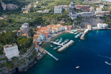 İtalya 'nın Sorrento kentinin yüksek açılı panoramasında küçük bir marina ve dramatik uçurumlar etrafında hareketli evler yükseliyor. Sahnede turkuaz Akdeniz suyu, yemyeşil yemyeşil ve canlı bir kıyı atmosferi yer alıyor..