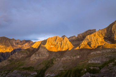 Gece geç saatlerde güneş, engebeli dağ tepeleri ve Pireneler 'deki kayalık yamaçlar boyunca parlak bir altın parıltı saçar. Karamsar bir gökyüzü altında, ışığın ve gölgenin dramatik zıtlıkları olur..