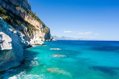 Sardunya 'nın engebeli Cala Goloritze kıyı şeridi boyunca kristal berrak turkuaz suyun üzerinde dik kireçtaşı kayalıklar yükselir. Parlak güneş ışığı ve parlak mavi bir gökyüzü çarpıcı, sakin bir Akdeniz manzarası yaratır..