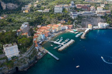 İtalya 'nın Sorrento kentinin yüksek açılı panoramasında küçük bir marina ve dramatik uçurumlar etrafında hareketli evler yükseliyor. Sahnede turkuaz Akdeniz suyu, yemyeşil yemyeşil ve canlı bir kıyı atmosferi yer alıyor..