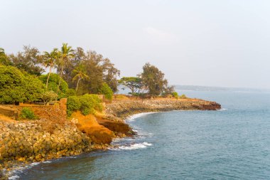 Yeşiller yemyeşil ağaçlar ve tarihi taş tahkimatları Hindistan 'ın Kannur kenti yakınlarındaki Arap Denizi' nin sakin mavi sularına kayalık bir burnu oluşturur. Sıcak güneş ışığı kayaların, yaprakların ve kıyı manzarasının zengin dokusunu vurgular.