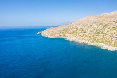 Wide aerial view of a rugged, sunlit rocky headland jutting into the deep blue Mediterranean waters near Preveli, Crete. The coastline features arid slopes with sparse greenery under a clear sky, creating a tranquil and open atmosphere.