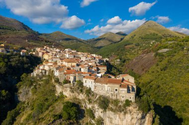İtalya 'nın Tortora kentinin yüksek manzarası, terakotta çatıları olan güneşli taş evler, çarpıcı bir uçurum kenarına yığılmış, yemyeşil tepeler ve dağınık bulutlu mavi gökyüzünün altındaki engebeli dağlar..