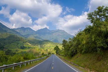 Kuzey Vietnam 'daki dramatik dağ zirvelerinin altındaki yemyeşil tepeler ve vadiler boyunca dümdüz asfalt yol esiyor. Parlak gün ışığı ve dağınık bulutlar canlı, davetkar bir manzara yaratır..