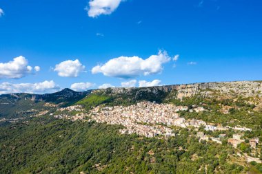 İtalya 'nın Sardunya kentindeki dramatik kireçtaşı kayalıklarının altındaki sık ağaçlı bir yamaçta yer alan Baunei köyünün geniş bir manzarası. Parlak güneş ışığı ve dağınık kümülüs bulutları canlı bir Akdeniz atmosferi yaratır..