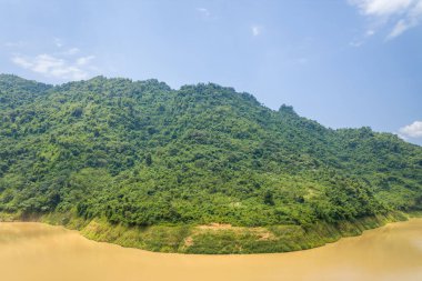 Yoğun ormanlık bir dağ Kuzey Vietnam 'da geniş ve çamurlu bir nehrin üzerinde kıvrılarak yükseliyor. Parlak gün ışığı canlı yeşilliği vurgular ve suda yumuşak yansımalar yaratır..