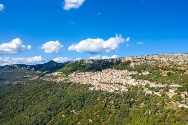 Panoramik bir hava fotoğrafı, İtalya 'nın Sardunya kentindeki engebeli kireçtaşı kayalıklarına yuva yapmış Baunei köyünü yakalıyor. Sahnede yemyeşil tepeler, dağınık bulutlar ve Akdeniz manzarasını aydınlatan canlı mavi gökyüzü yer alıyor..