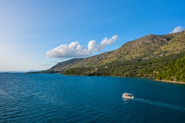 Yunanistan 'ın Epirus kentindeki Plataria' nın yeşil tepeleri ve engebeli kıyı şeridi yakınlarındaki berrak mavi sularda küçük bir tekne hareket ediyor. Sahnede parlak gün ışığı, dağınık bulutlar ve Akdeniz manzarasına karşı durgun İyon Denizi yer alıyor..