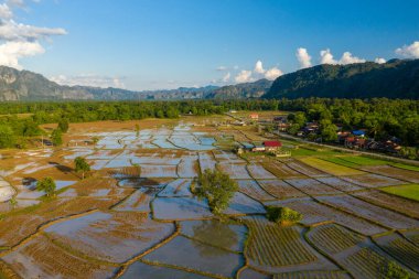 Sel basmış pirinç tarlalarının gökyüzünü yansıtan hava manzarası, arka planda kireçtaşı tepeleri olan küçük bir köy ve yoğun yeşil ormanla çevrili. Sıcak akşam ışığı geometrik desenleri ve sakin kırsal atmosferi vurgular.