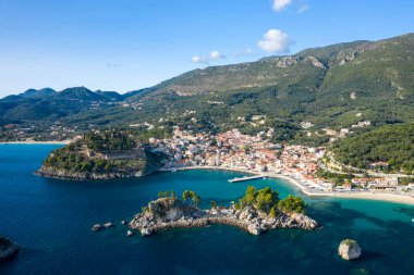 Wide aerial view of Parga in Epirus, Greece, displaying a vibrant coastal village with red-roofed houses, a historic hilltop fortress, and rocky islets surrounded by turquoise waters. The Mediterranean landscape is framed by densely forested hills