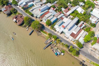 Hava görüntüsü, Can Tho, Mekong Delta, Vietnam 'daki Cai Rang' ın nehir kıyısında uzanan metal çatılı evler ve yemyeşil ağaçlar yakalıyor. Çamurlu su boyunca birkaç uzun tekne demirlemiş, kıyı şeridinde gölgeler bırakan palmiyeler var.