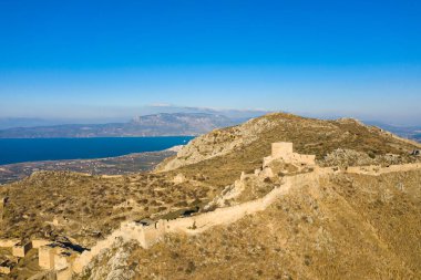 Kırılan taş duvarlar ve antik kalıntılar, Yunanistan 'da berrak bir gökyüzü altında derin mavi körfez ve uzak dağların süpüren manzaralarıyla, Akrokorinth' in güneşli yamaçları boyunca uzanır..