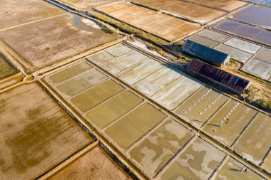 Drone perspektifi, Kamboçya 'nın Marais tuzlu su depolarındaki tuz buharlaştırma havuzlarının geometrik düzenini ve iki uzun yıpranmış depoyu yakalar. Sahnede toprak dokular, yamalı alanlar ve sıcak güneş ışığı altında ince yansımalar yer alıyor..