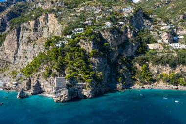 İtalya 'nın Positano kenti yakınlarındaki turkuaz suların üzerindeki engebeli uçurumlarda tarihi bir taş gözetleme kulesi bulunuyor. Dağılmış yamaçlardaki villalar ve yemyeşil Akdeniz bitki örtüsü parlak güneş ışığı altındaki dramatik kıyı manzarası..