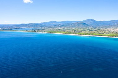 Sardunya 'nın Lotzorai kenti yakınlarındaki yemyeşil kıyı şeridi ve uzak dağlarla buluşan canlı mavi Akdeniz sularının berrak güneşli bir gökyüzü altında geniş bir hava manzarası. Sahne açık, aydınlık ve sakin. Sudaki hafif eğimlerle.
