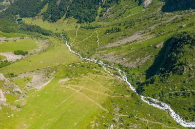 İnsansız hava aracı, Pireneler 'deki engebeli yamaçlar ve dağınık ağaçlarla çevrili yeşil bir vadide gezinen dar bir dağ nehrine bakıyor. Parlak yaz güneşi altında..