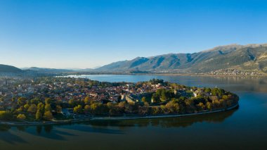 Yumuşak güneş ışığı, Ioannina Gölü 'nün sakin suları ve Pindus dağlarının engebeli yamaçlarıyla çevrili tarihi yarımadasını aydınlatır. Görüntüde kırmızı kiremitli çatılar, ortaçağ duvarları ve yemyeşil alanlar yer alıyor.
