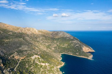 Yunanistan 'ın Magne kentindeki tenha bir kayalık koya ve derin mavi Akdeniz sularına inen engebeli tepelerin havadan görünüşü. Güneş ışığı yumuşak bulutlarla dağılmış parlak bir gökyüzünün altındaki desenli manzarayı vurgular..