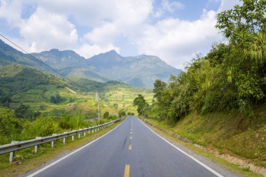 Paved yolu yemyeşil tepeler ve yoğun yeşillikler arasındaki mesafeye uzanır. Kuzey Vietnam 'da parlak bir gökyüzünün altında dramatik dağ zirveleri yükselir. Güneş ışığı canlı manzarayı vurgular ve açık bir macera hissi yaratır..