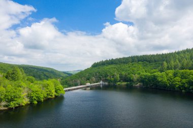 Yoğun yeşil ormanlarla çevrili sakin bir gölün yüksek açılı perspektifi, uzun bir baraj ve parlak mavi gökyüzünün altında dağınık bulutlarla kaplı Chaumecon gölünde küçük bir bina..
