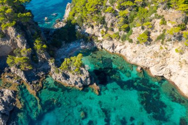 Engebeli uçurumlarla ve İyon Denizi kıyısındaki yoğun çam ağaçlarıyla çevrili berrak bir turkuaz göl perspektifi. Güneş ışığı canlı sualtı dokularını ve kayalık kıyı şeridini vurguluyor ve canlı bir Akdeniz atmosferi yaratıyor..