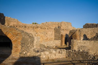 Ilık güneş ışığı, İtalya 'nın Pompeii kentindeki arkeolojik kamaralarda yıpranmış volkanik taşlar ve tuğla kemerleri aydınlatıyor. Tarihi taş ustası, parlak mavi gökyüzünün altında pürüzlü dokuları ve katmanlı yapıları ortaya çıkarıyor..