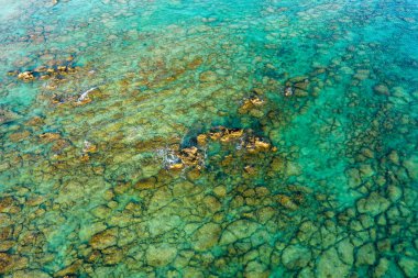 Girit yakınlarındaki sığ bir Akdeniz kıyı şeridinin yukarıdan aşağı görüntüsü, berrak turkuaz suyun altındaki dokulu kayaları ortaya çıkarıyor. Güneş ışığı sualtı desenlerini ve canlı kıyı renklerini vurgular.